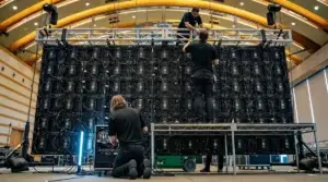 Behind a flown LED wall during corporate event build, showing vertical bundles of CAT5 ethernet/fiber signal cables and 12 AWG black power feeder cables running down the back of LED cabinets, ground-supported truss with chain motors at the top corners, two ProCore crew members in solid black polos mid-build with one on a Genie scissor lift trimming the wall and one kneeling at the floor signal rack