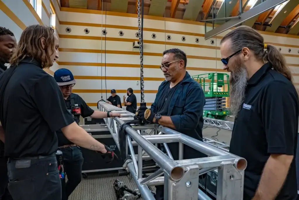 Five ProCore AV crew members in matching black polos assembling a stack of aluminum truss inside a wood-ceiling Los Angeles venue during corporate event load-in, hands on the truss segments mid-build with road cases and a scissor lift visible in the background