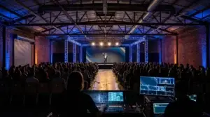 Corporate keynote in progress inside a converted Los Angeles warehouse, with a speaker on stage in front of a large LED wall framed by ground-supported truss with cobalt blue and amber stage wash, audience seated theater-style facing the stage, exposed red brick walls and steel structural beams visible, and a production tech table with multiview monitor visible at the back of the room facing the stage